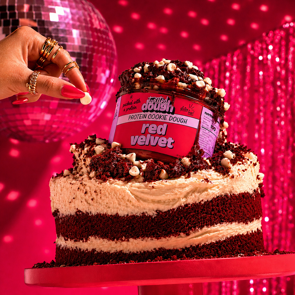 Red velvet cake with a container of Twisted Dough red velvet protein cookie dough on top, against a disco ball and curtain background.